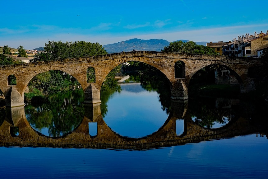 A beautiful artistic photo of a bridge over  a canal reflecting the blue sky with mountains in the background on a clear day